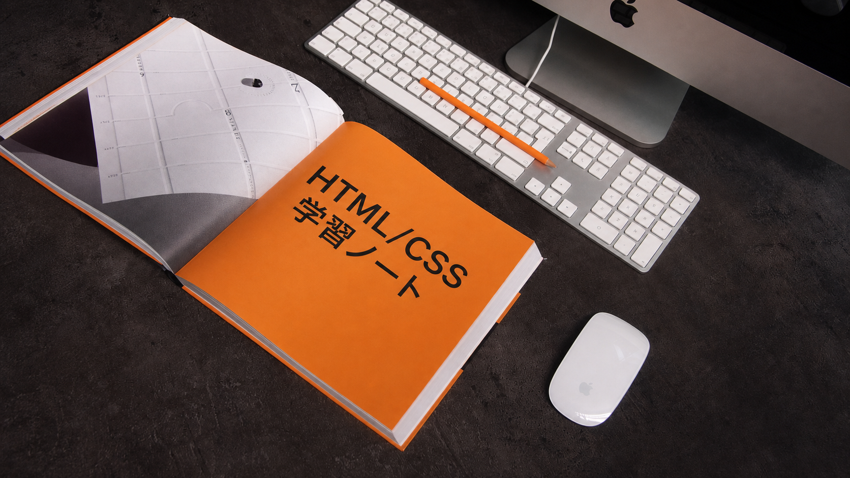 パソコンの横でHTMLとCSSの学習ノートを開いている写真